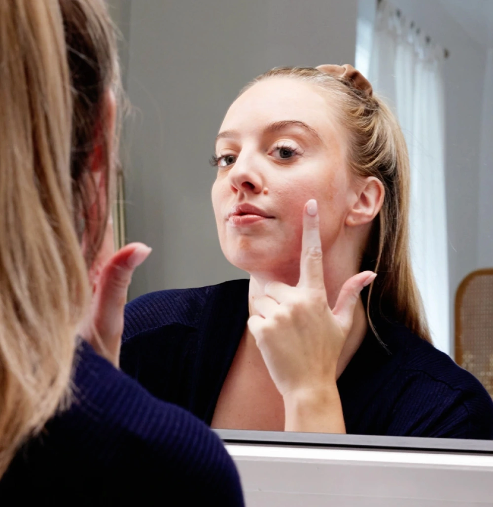 A young woman with vitiligo confidently applying treatment in the mirror. She is holding up her pointer finger applying the treatment to her cheek area.  